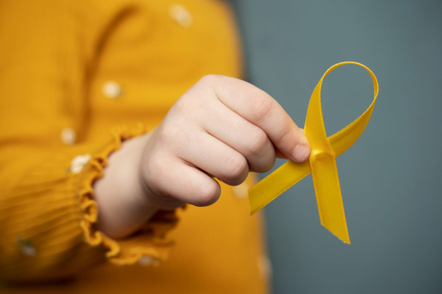 A close-up shot of a child's hand holding a yellow ribbon loop, a symbol for childhood cancer awareness. The child is wearing a textured mustard-yellow long-sleeved shirt against a soft, dark background.