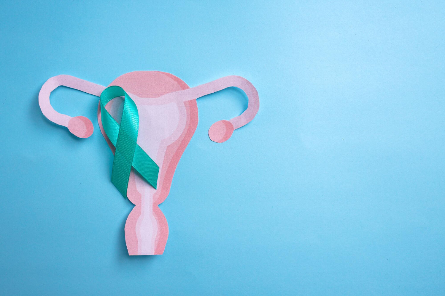 Top-down view of a light pink paper illustration representing the uterus, fallopian tubes, and ovaries, placed on a bright blue backdrop. A folded teal awareness ribbon rests on the left side of the paper cutout.
