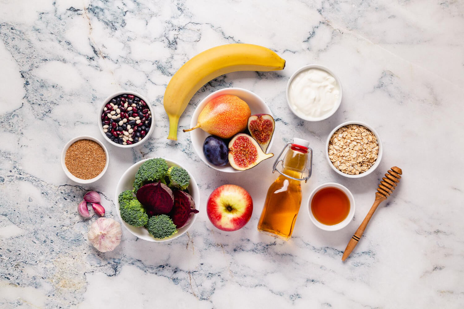 A top-down view on a light marble surface displaying various gut-healthy foods, including a yellow banana, a bowl with a pear, cut figs, and a plum, a red apple, a bowl of broccoli florets and sliced beet, a bowl of mixed beans, a bowl of ground flaxseed or grain, garlic cloves, a small bowl of yogurt, a bowl of rolled oats, a glass bottle of apple cider vinegar, and a small bowl of honey with a wooden dipper.