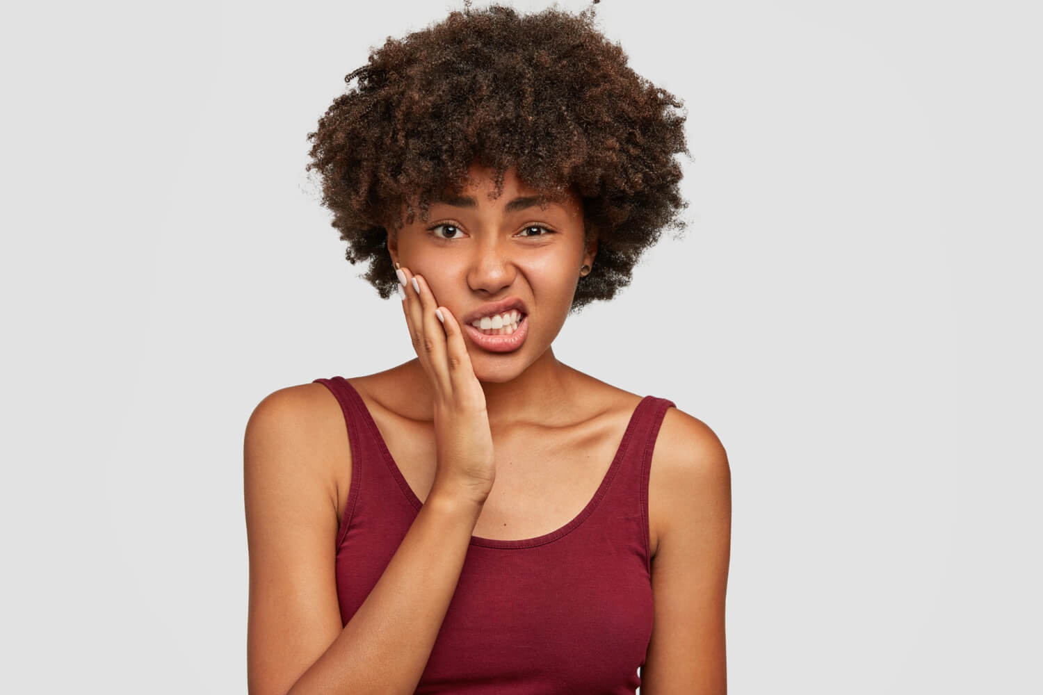 A young Black woman with an afro holding her cheek in pain while wincing against a plain white background.