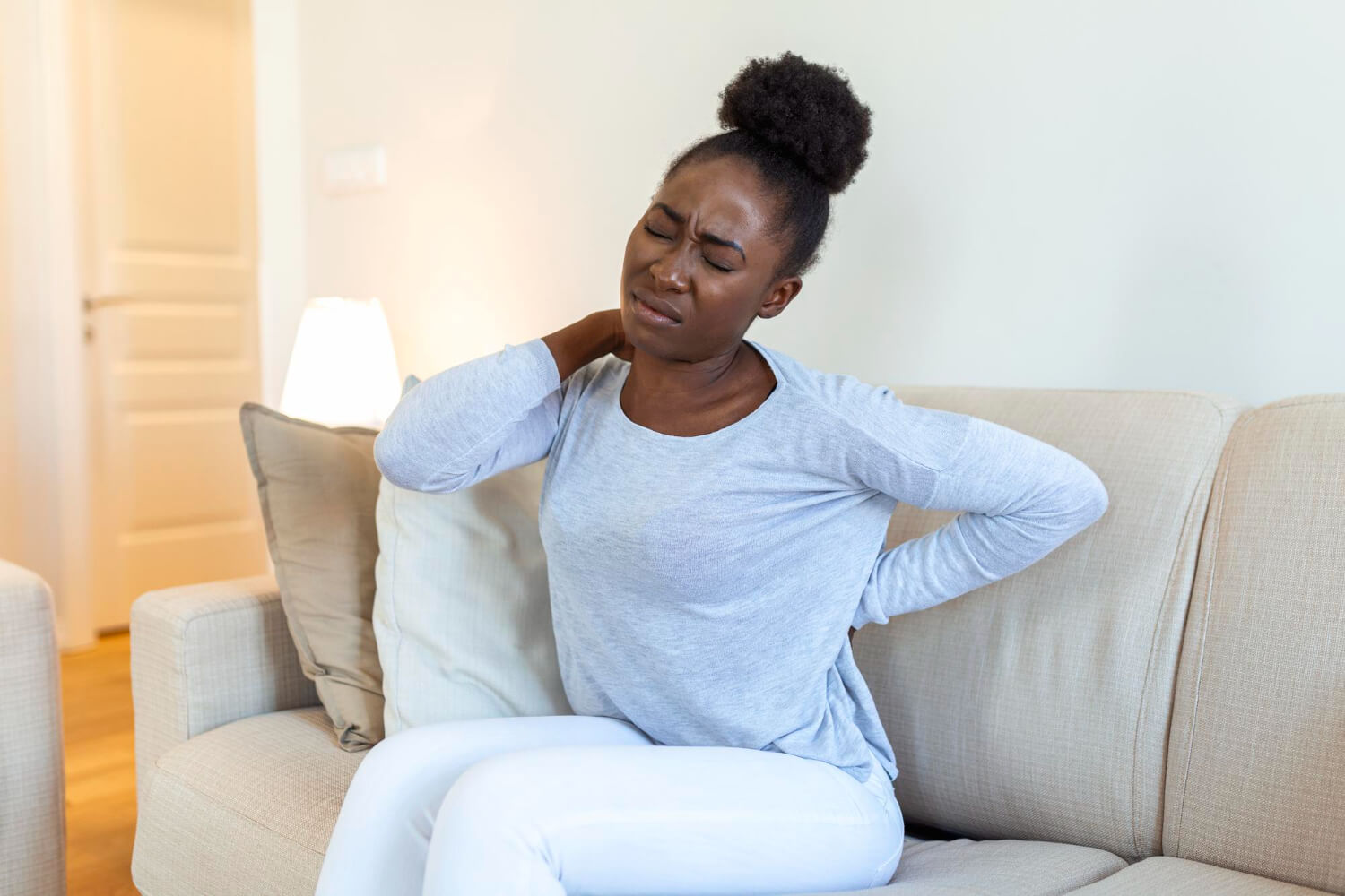 A woman sitting on a couch with her eyes closed in pain, holding the back of her neck with one hand and her lower back with the other.