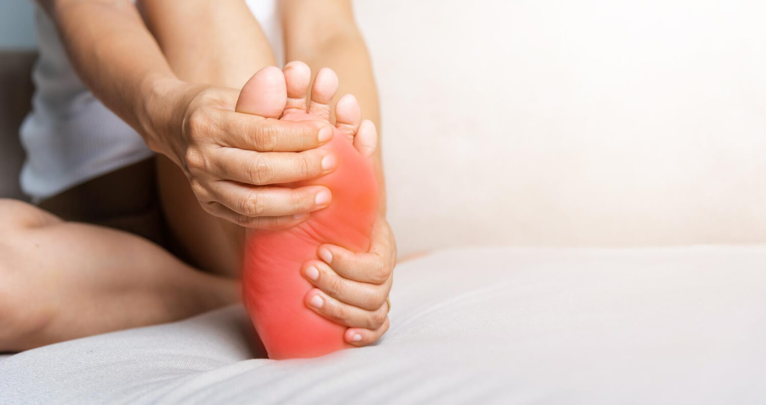 A person holding the sole of their bare foot with both hands, with a red highlighted overlay covering the heel and sole area to illustrate pain or inflammation.