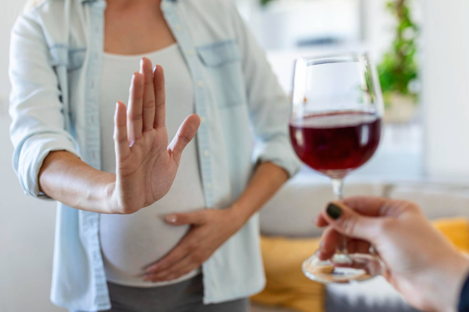 Pregnant woman holding up her hand to refuse a glass of red wine offered by another person.