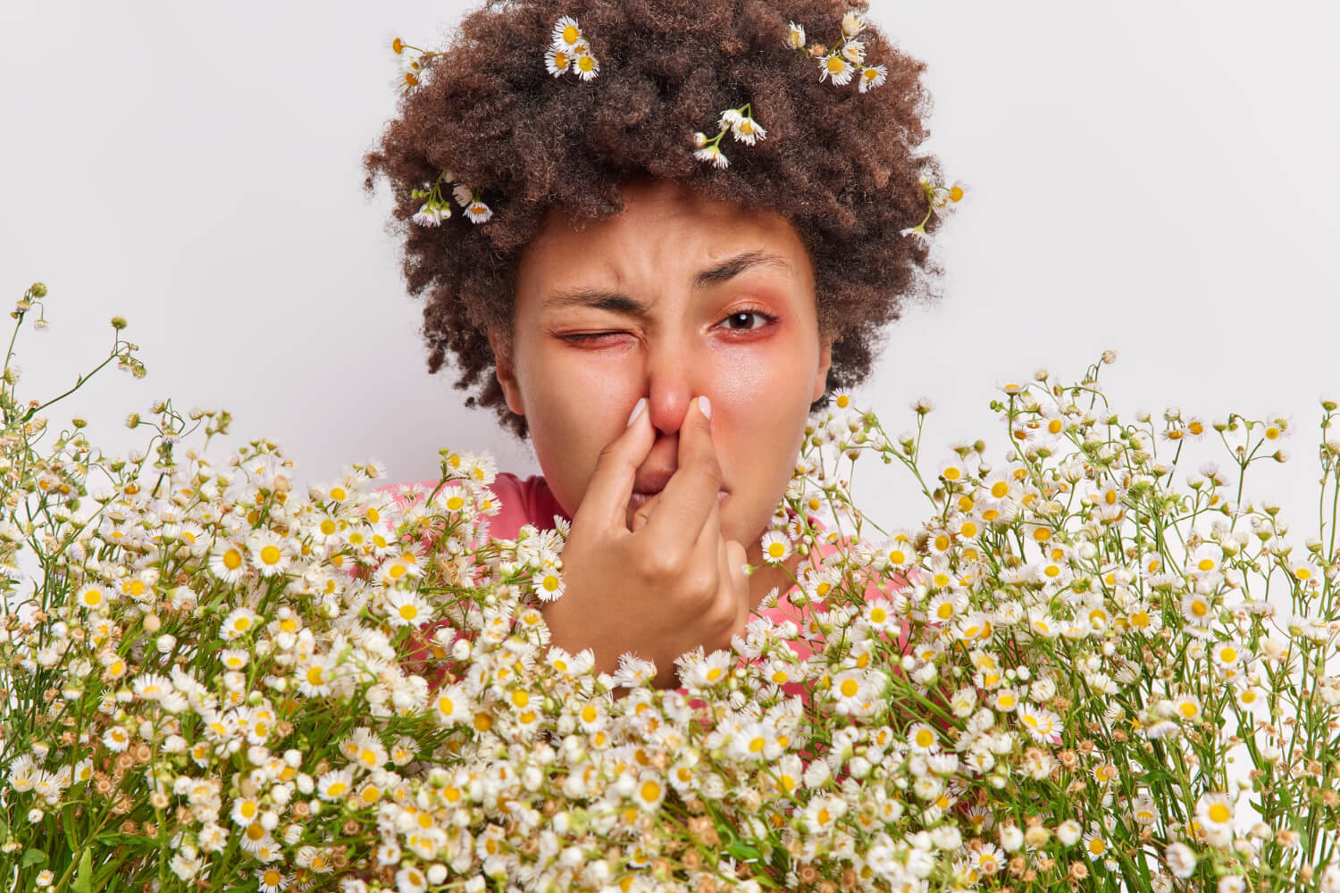 A woman surrounded by small white and yellow daisy-like flowers pinching her nose closed while winking one reddened, irritated eye.
