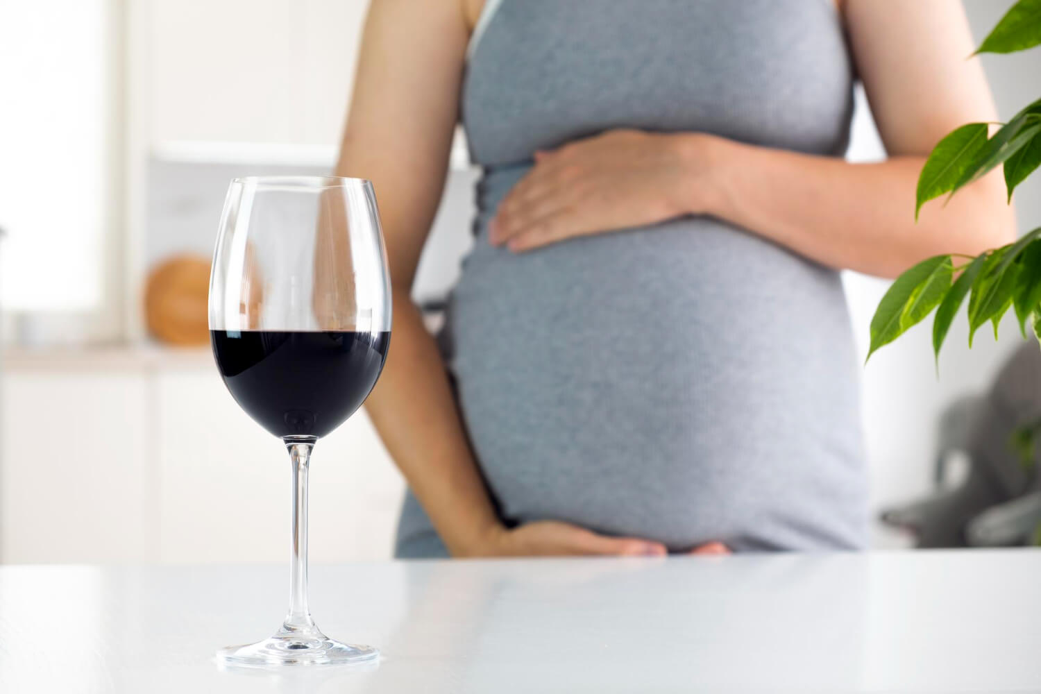 A glass of red wine sitting on a white table in the foreground with a pregnant woman standing blurred in the background.
