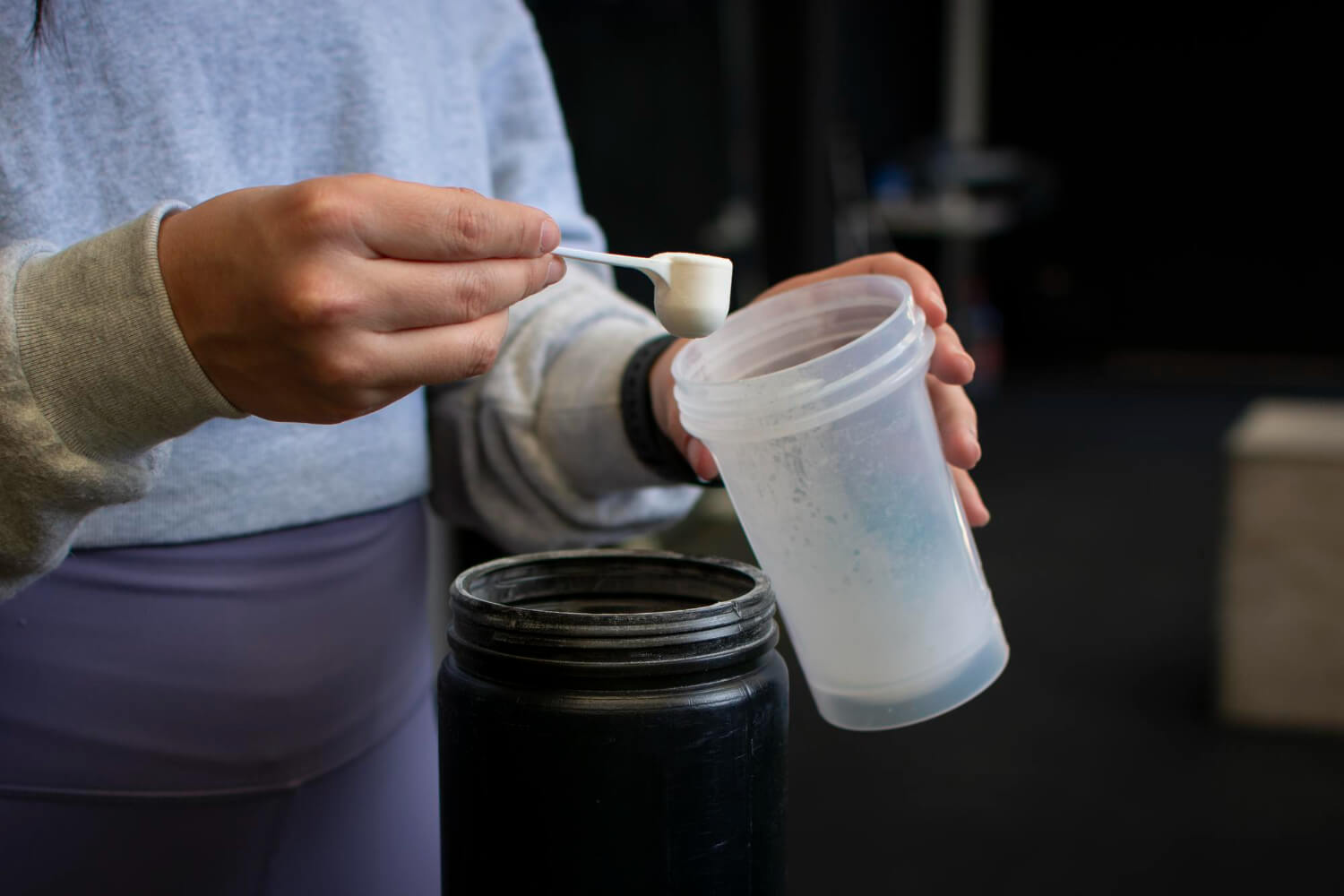 A person holding a scoop filled with white powder over an open shaker bottle, with a black powder tub underneath.