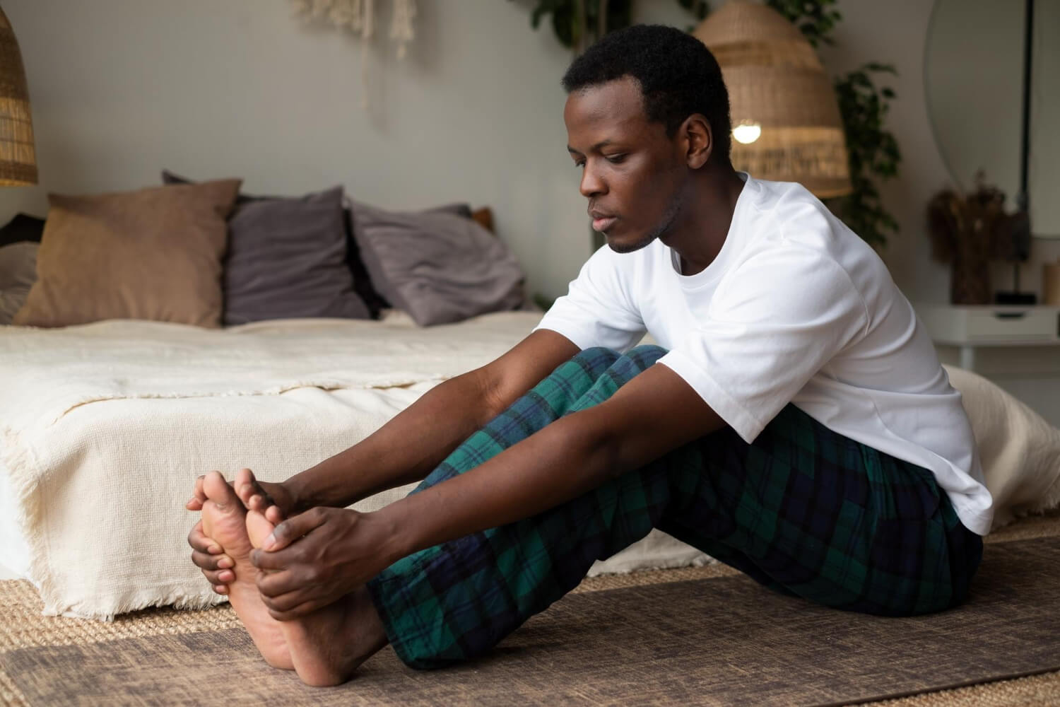 A man in a white t-shirt and green plaid pajama pants sitting on the floor next to a bed, leaning forward to hold both of his feet with his hands.