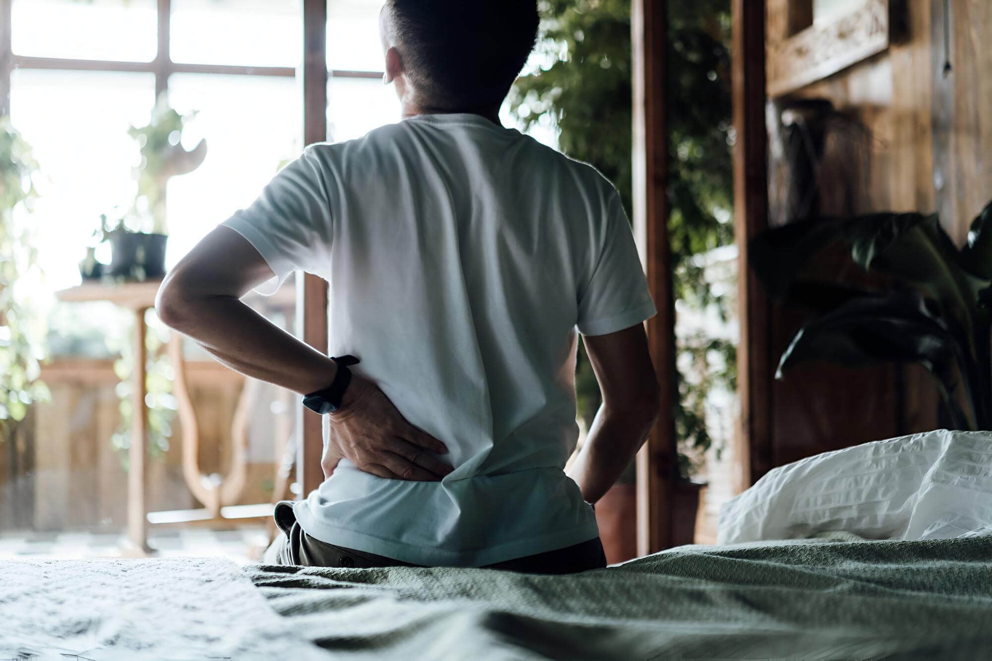 Rear-view of a person sitting on the edge of a bed, wearing a white t-shirt and holding their lower back in pain.