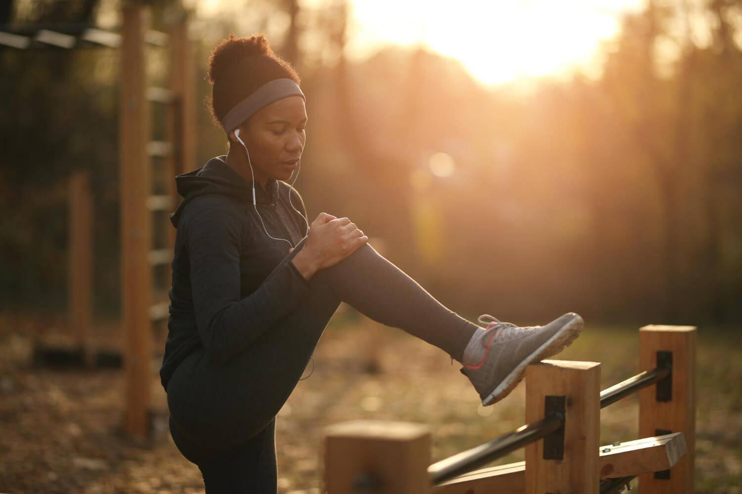 A woman wearing a dark long-sleeve workout top, leggings, a headband, and wired earphones stretches outdoors in an exercise park. She rests one foot on a wooden post while holding her raised knee, bathed in warm, golden sunlight.