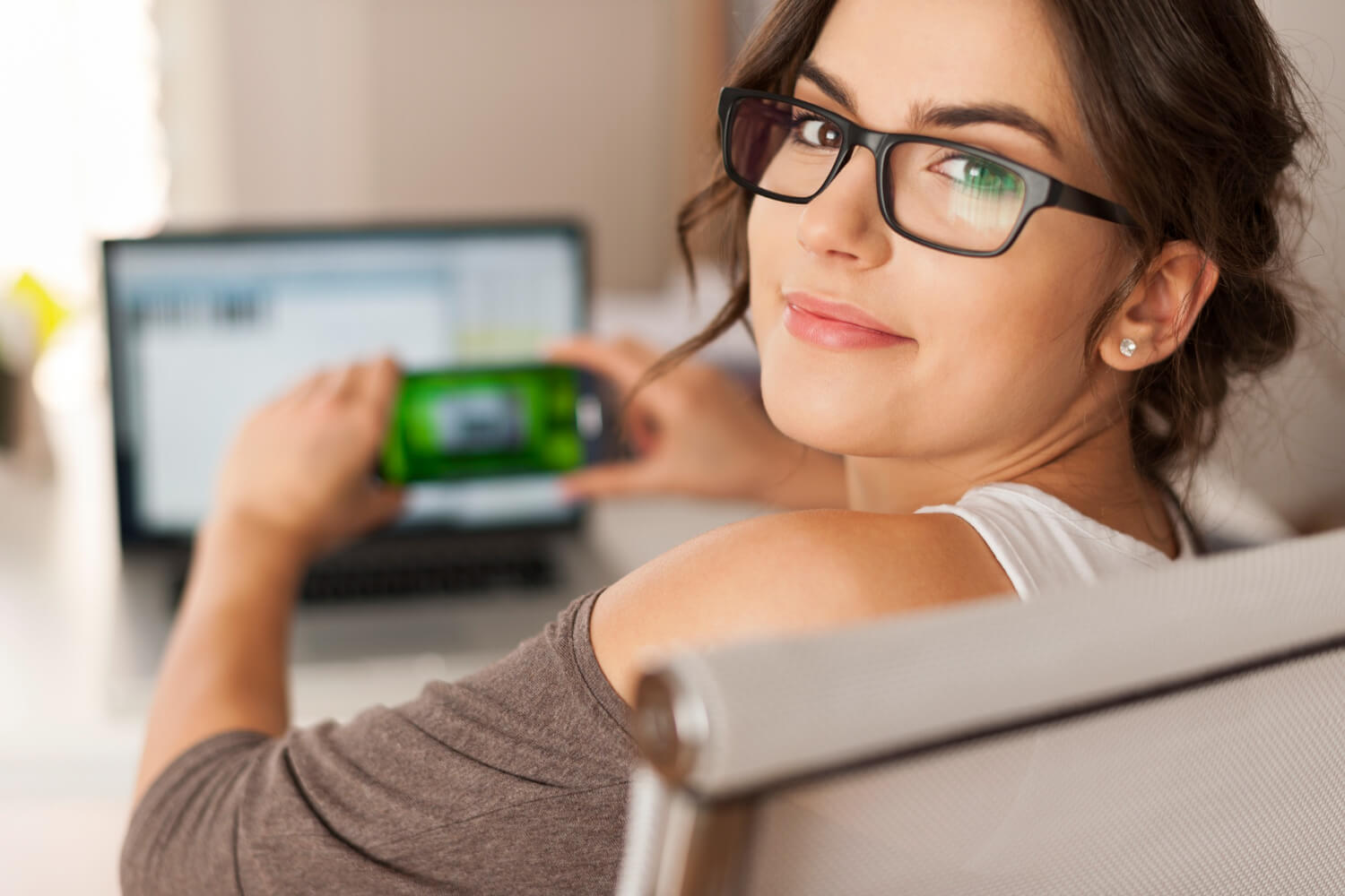 A woman wearing black-rimmed glasses looks back over her shoulder with a smile while holding a smartphone in front of a blurred laptop screen.