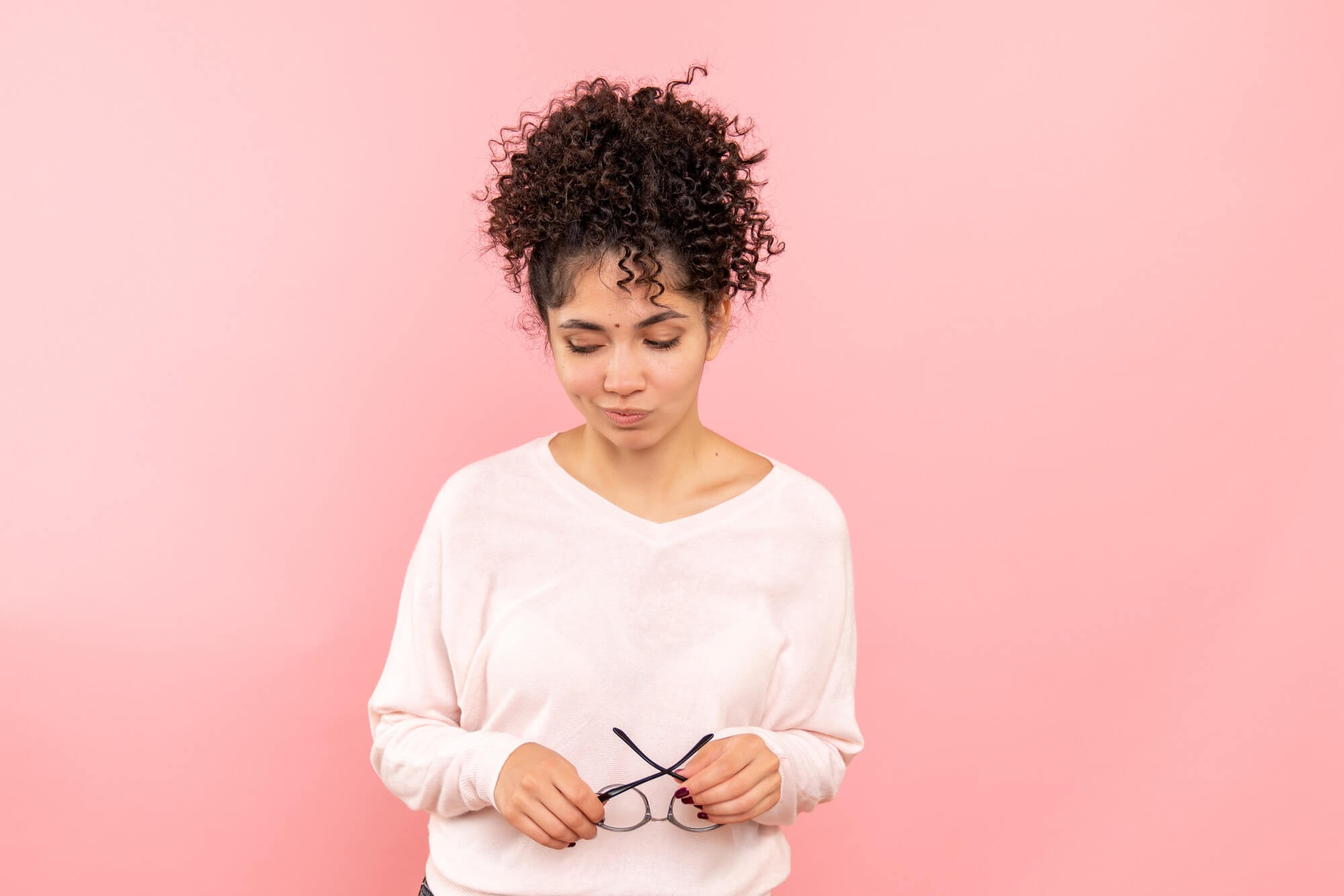 A young woman with curly hair pulled up into a high bun standing against a solid pink background, looking down thoughtfully while holding a pair of black-rimmed eyeglasses with both hands.
