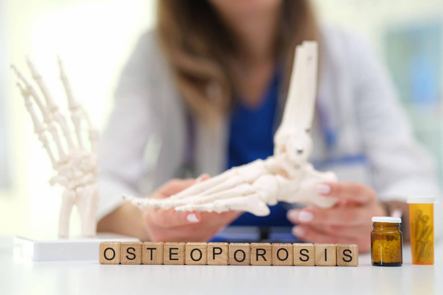 A healthcare professional in a lab coat holding a foot and ankle anatomical model, with wooden letter blocks spelling "OSTEOPOROSIS" and medicine bottles arranged on the desk in front.