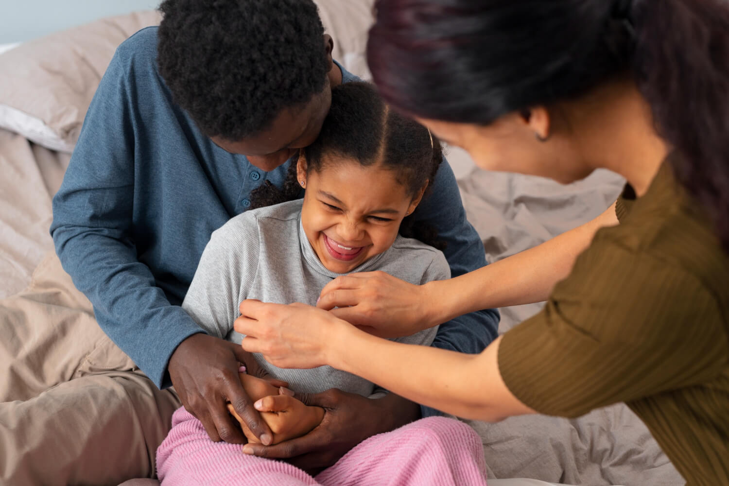 A young girl sitting between her parents on a bed, laughing playfully as her mother tickles her while her father holds her close from behind.